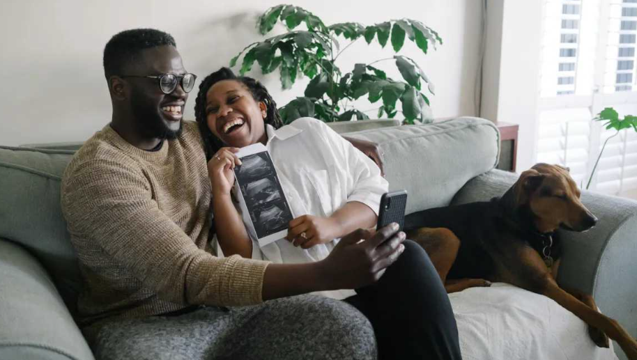 Un couple heureux partage un selfie d'annonce de grossesse à la maison, tenant des images d'échographie, parfait pour illustrer annonce grossesse facebook babylist